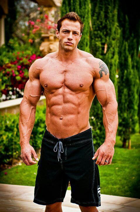 a muscular man with a six pack and a tanned body stands outdoors in a pair of black shorts. he's looking at the camera with a slight smile.  he looks fit and healthy, confident and strong. he's wearing a black and white pair of shorts.
