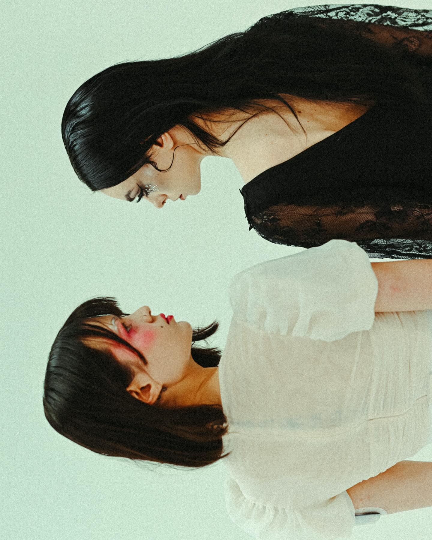 the image shows two women positioned almost mirroring each other. one woman is wearing a black, lacy, somewhat sheer dress; the other a white, sheer, puffsleeved top.  both women have dark hair and pale skin with subtle makeup; the woman in black has slightly more noticeable makeup. they are close together, with their faces slightly angled towards each other in what appears to be an intimate or intense confrontation. the background is a solid light green, keeping the focus completely on the models and their outfits. the overall mood is artistic, elegant and subtly mysterious.