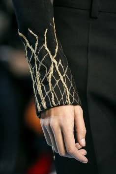a closeup shot of a person's hand wearing a black sleeve with gold embroidery. the sleeve is a unique design, with the embroidery forming a pattern that resembles branches or vines. the hand is posed to show off the detail of the sleeve and the intricate embroidery.