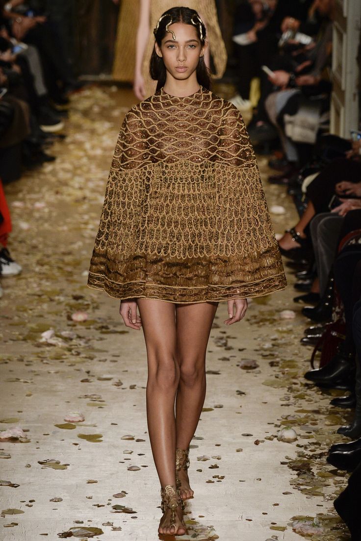 a model walks the runway in a gold dress with a capelike top, accessorized with a gold headpiece and sandals. the audience watches the fashion show. the floor is decorated with what appears to be petals or leaves.