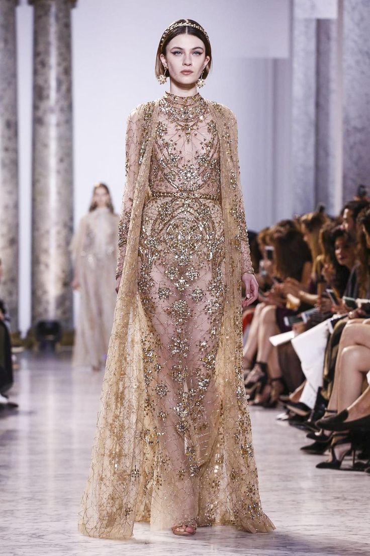 a model walks the runway wearing a stunning, floorlength gold and beige gown. the dress is heavily embellished with sparkles and intricate detailing, creating a luxurious and elegant look.  the dress features long sleeves and a capelike overlay. the model has a bob hairstyle and is wearing a gold headband and subtle jewelry. the background shows the audience watching the fashion show. the overall atmosphere is one of high fashion and sophistication.