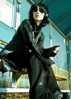 a young woman in black clothing and headphones sits in a bus stop, waiting.  her eyes are fixed on something in the distance, and her expression is serious. the photo is taken in a low light and has a dark, moody feel to it.  the lighting also casts shadows, making the photo more dramatic.
