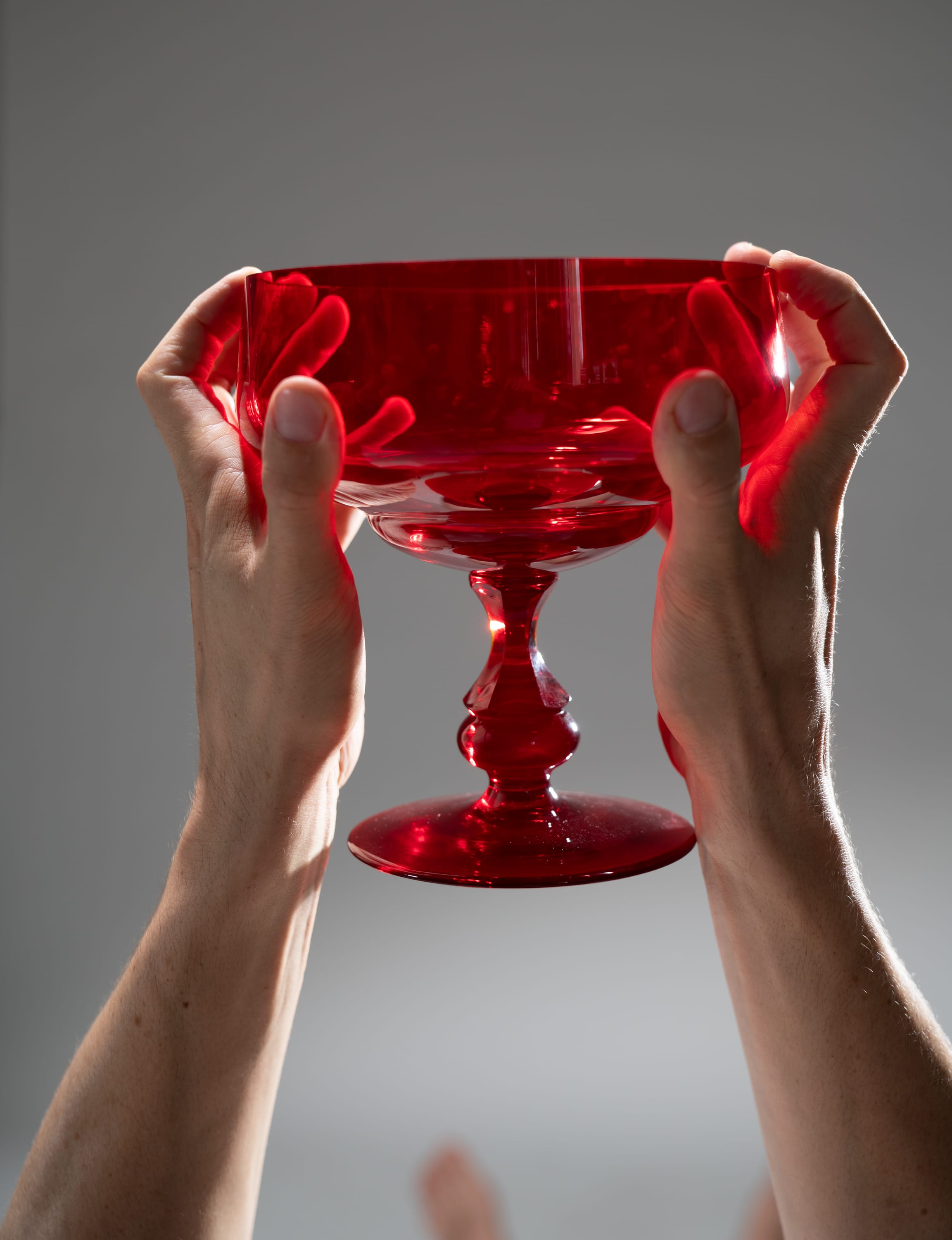 a closeup shot of a person holding a red, stemmed glass goblet. the goblet has a round bowl and a tall, thin stem. the glass is transparent, allowing light to pass through and create a reflection. the background is a neutral gray, which highlights the vibrant red color of the glass. the person's hands are positioned on either side of the bowl, securely holding the goblet.