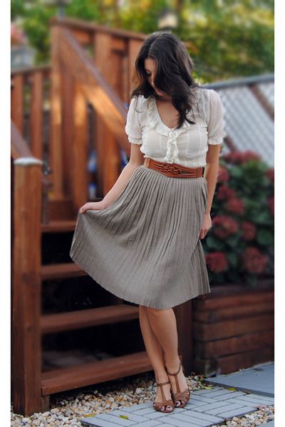 a young woman in a white shirt and a brown pleated skirt poses on some wooden steps outdoors. she is wearing brown sandals and has long brown hair. she is looking down at her feet.