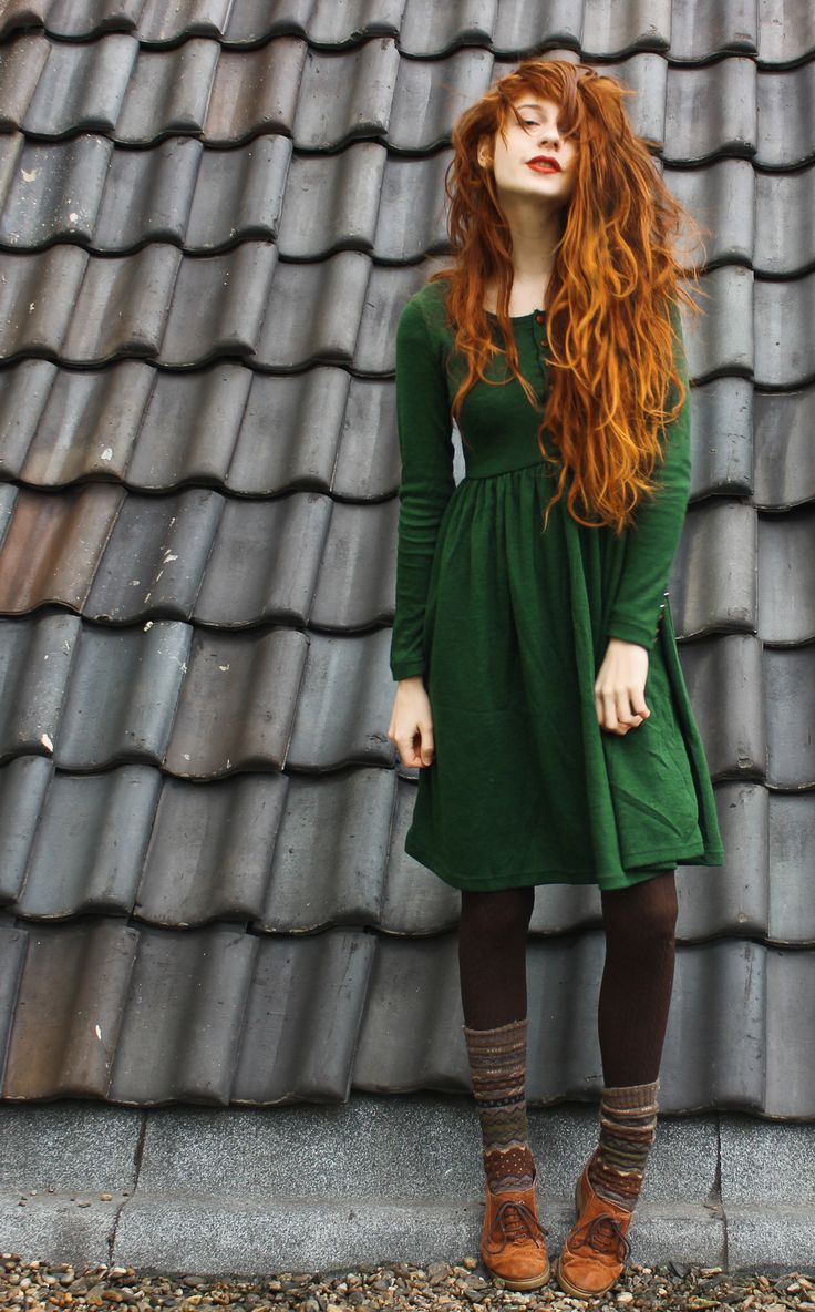 a young woman with long red hair stands on a rooftop with grey tiles. she is wearing a longsleeved, green dress, brown tights with patterned tops, and brown laceup boots. she is posed with her head tilted back and eyes closed. the background is a wall of grey tiles, and the lighting is overcast.