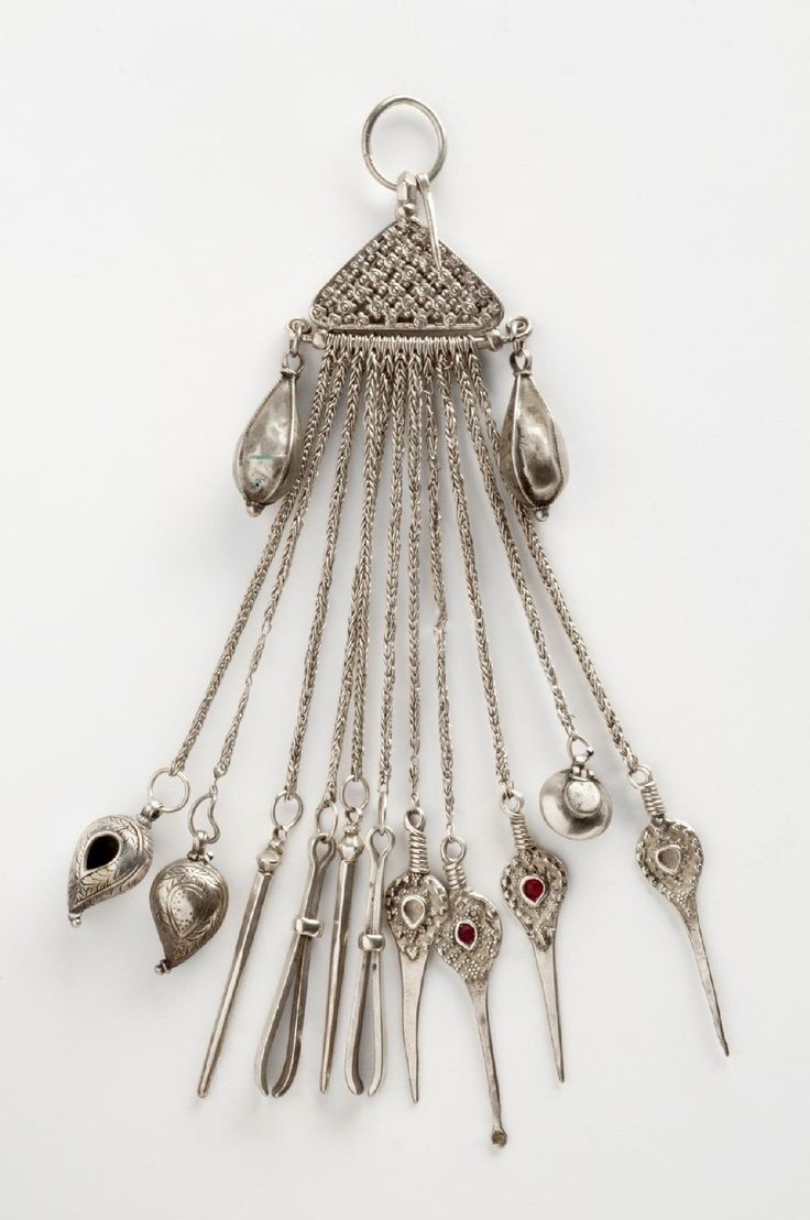 the image displays an antique silver pendant featuring an intricate design. the pendant has a triangular top with a ring for attachment. dangling below are multiple chains with various silver charms and tools, including scissors, cosmetic tools, and decorative elements, showcasing fine metalwork and craftsmanship.