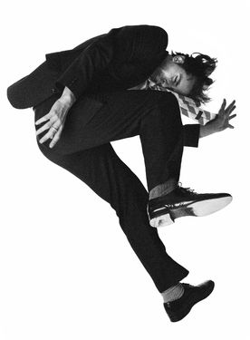 a black and white photo of a man dancing and midair in a suit and tie. he has one leg raised and his arm is stretched out.  the picture is taken midjump with the man facing his right.