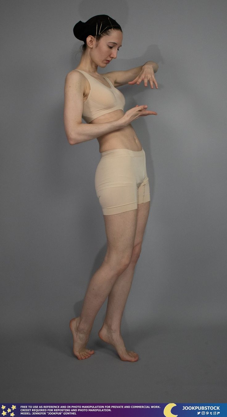 a young woman is posing in the studio, wearing a tank top and shorts. she has her feet slightly apart, and her hands are posed as if she is holding something. she is looking down at her hands.  her hair is pulled back in a bun and she is wearing a black hair pin. she is standing on a grey background.