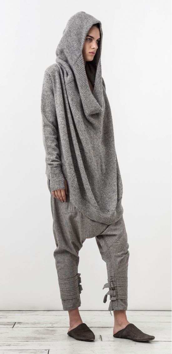 a full body shot of a woman dressed in gray loungewear, comprising of a hooded tunic sweater with drop crotch pants, and slippers. the woman is standing in a studio setting with a white backdrop.