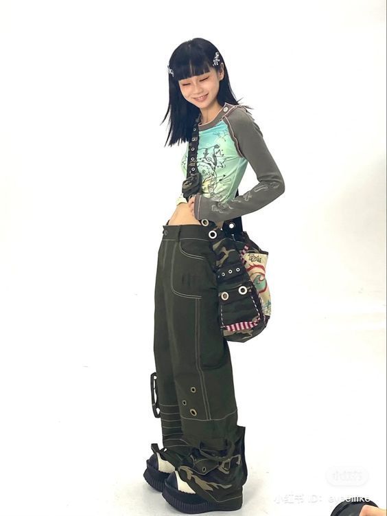 the image shows a young woman posing in a studio setting against a white background. she is modeling a y2kinspired outfit which includes baggy, army green cargo pants with stitching details, a pale green and brown graphic longsleeved top,  camouflage print platform boots and a patterned shoulder bag. her hair is styled with black bangs and small hair clips. her overall style is characterized by a mix of grunge, alternative, and streetwear elements.  the image is brightly lit and clearly shows the details of her clothing and accessories.