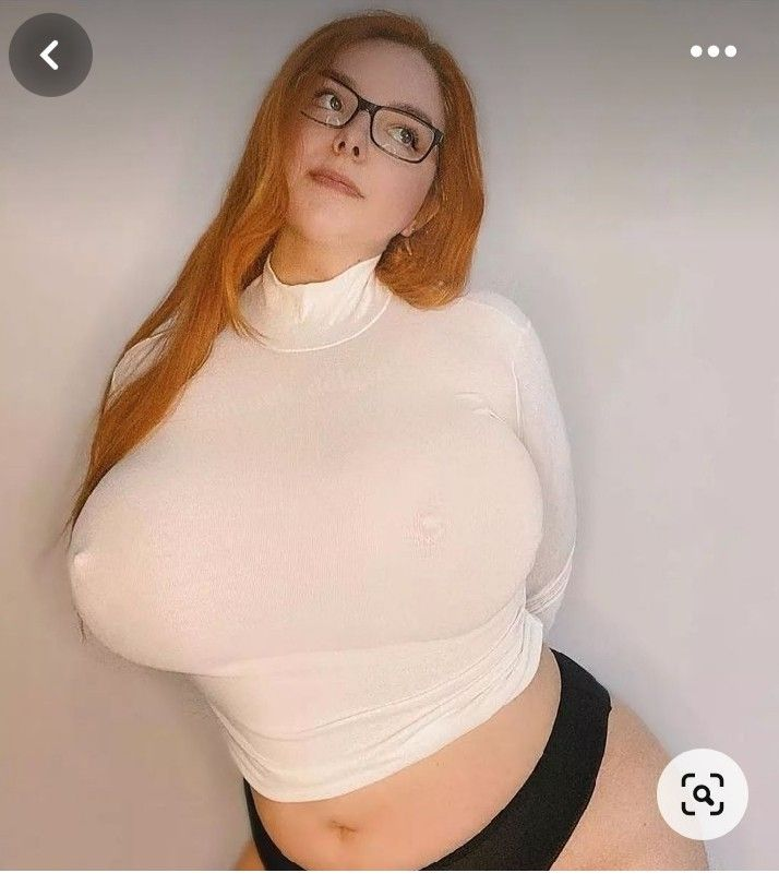 a woman with red hair and glasses is pictured from the chest up. she is wearing a white turtleneck and black underwear. she is looking up and to the left. her skin is pale.