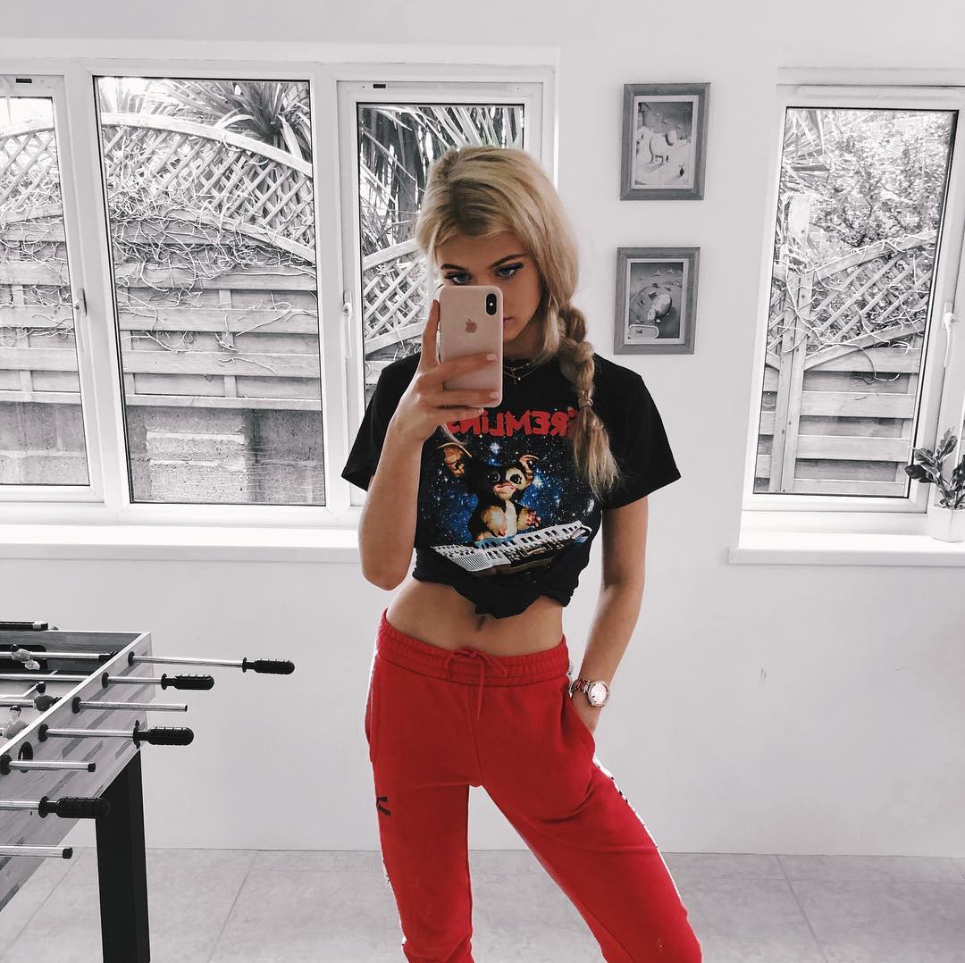 a woman with blonde hair in a braid is taking a selfie with her phone. she is wearing a black tshirt with a graphic design, red sweatpants, and a watch. she is standing in a room with windows, picture frames, and a foosball table. the room appears to be the interior of a house.