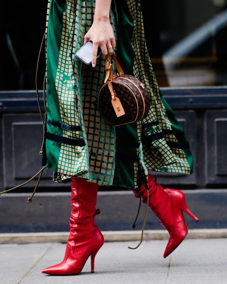 a woman is walking down the street, wearing a stylish green and gold patterned dress, red stiletto ankle boots, and a louis vuitton round handbag. she is holding a smartphone in her hand. the photo is taken from a midleg perspective, emphasizing the details of her outfit and accessories. the overall style is chic and modern. the setting appears to be an urban area.