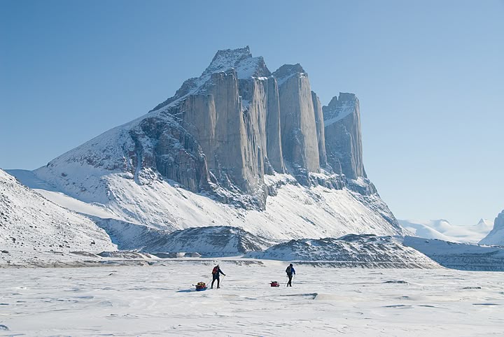 the image shows a snowcovered landscape with a large mountain range in the background. two people are pulling sleds across the snow in the foreground. the sky is clear and blue.