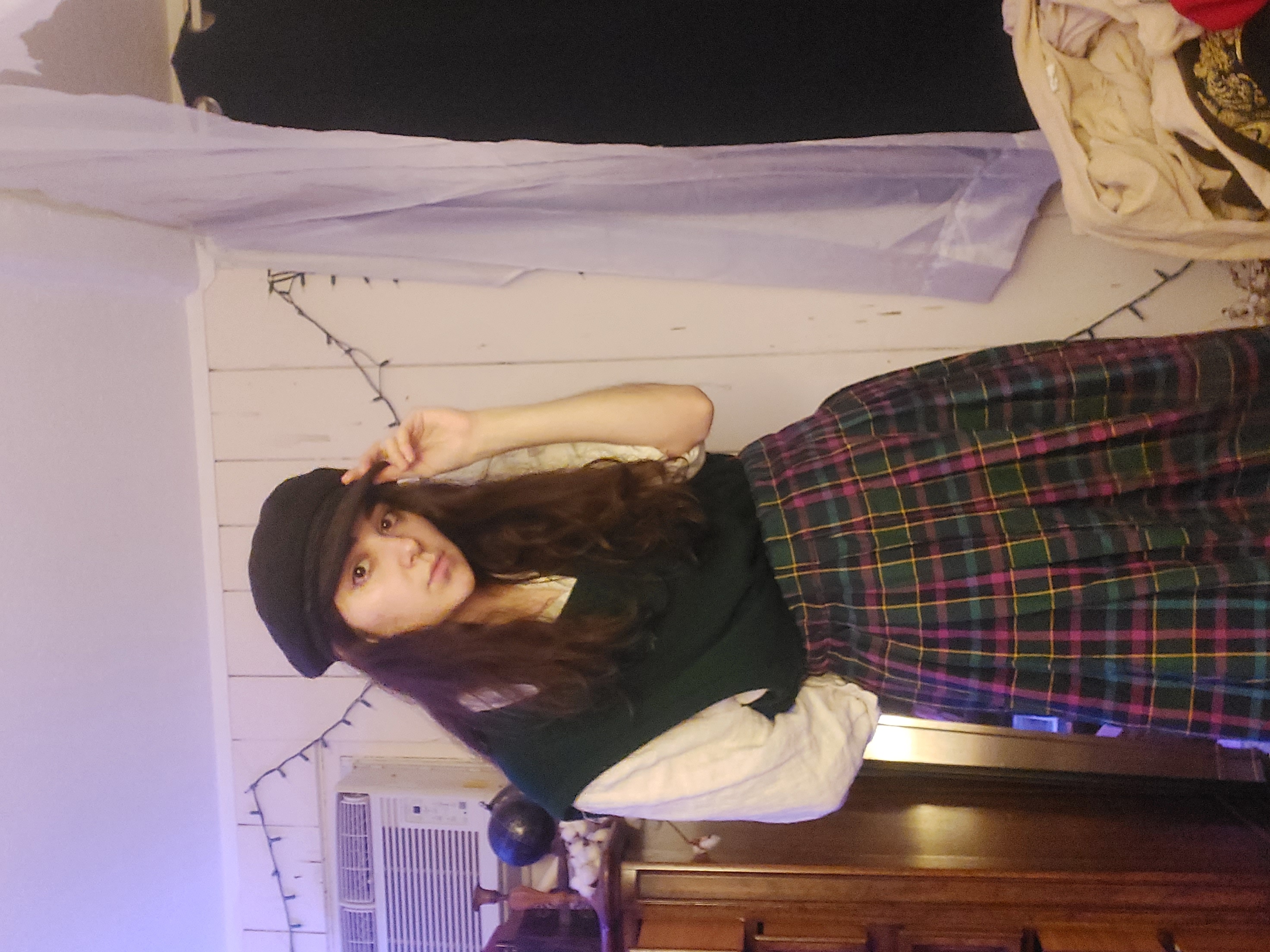 a young woman poses for a selfie in her bedroom. she's wearing a dark green sweater vest over a white longsleeved shirt, a colorful plaid pleated skirt, and a dark newsboy cap.  her long brown hair is loose. she is standing in front of a dresser and a clothes rack with items hanging on it. string lights are visible on the wall in the background.  the overall style is casual and slightly retro or vintage.
