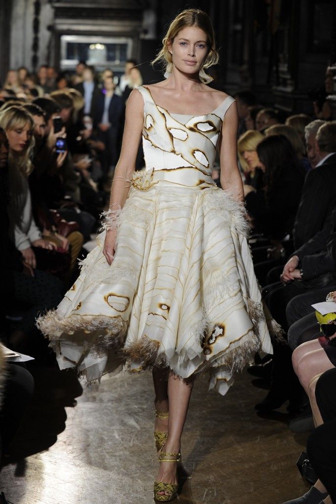 a fashion model walks down the runway in a white dress with brown detailing and a feather trim. the dress has a cinched waist and a full skirt. the model is wearing her hair in a sleek updo and has minimal makeup. the show is taking place in a dark room with a seated audience. the model is looking down towards the ground.