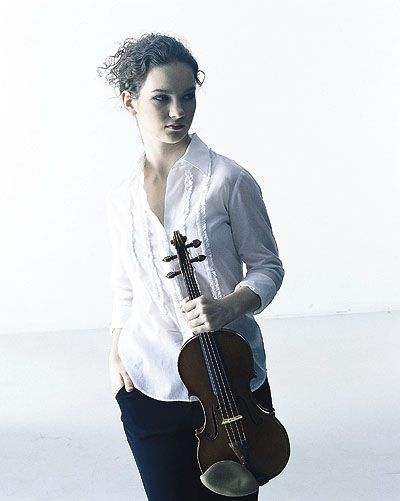 a young woman, formally dressed in a white buttondown shirt and black pants, stands in a white studio space. she holds a violin upright in her left hand, resting her other hand in her pocket.  her gaze is directed offcamera, creating a sense of contemplation or anticipation. the image's soft lighting and minimal backdrop draw focus to the musician and her instrument.