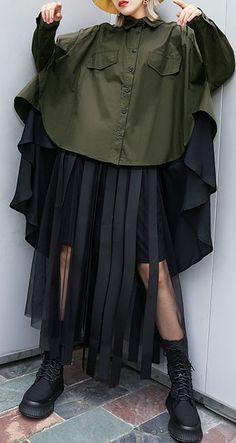 a woman is standing with her legs spread slightly apart, wearing an oversized green shirt that has a black mesh overlay. she's wearing a black skirt that has pleats, and black boots. she's looking slightly to the right of the camera. the background is plain white.