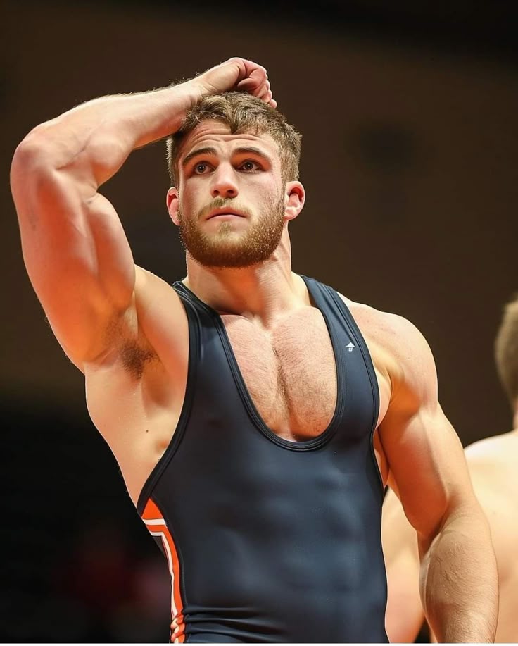 a muscular man wearing a wrestling singlet is standing with his hand on his head, looking up. he has a beard and an athletic physique.