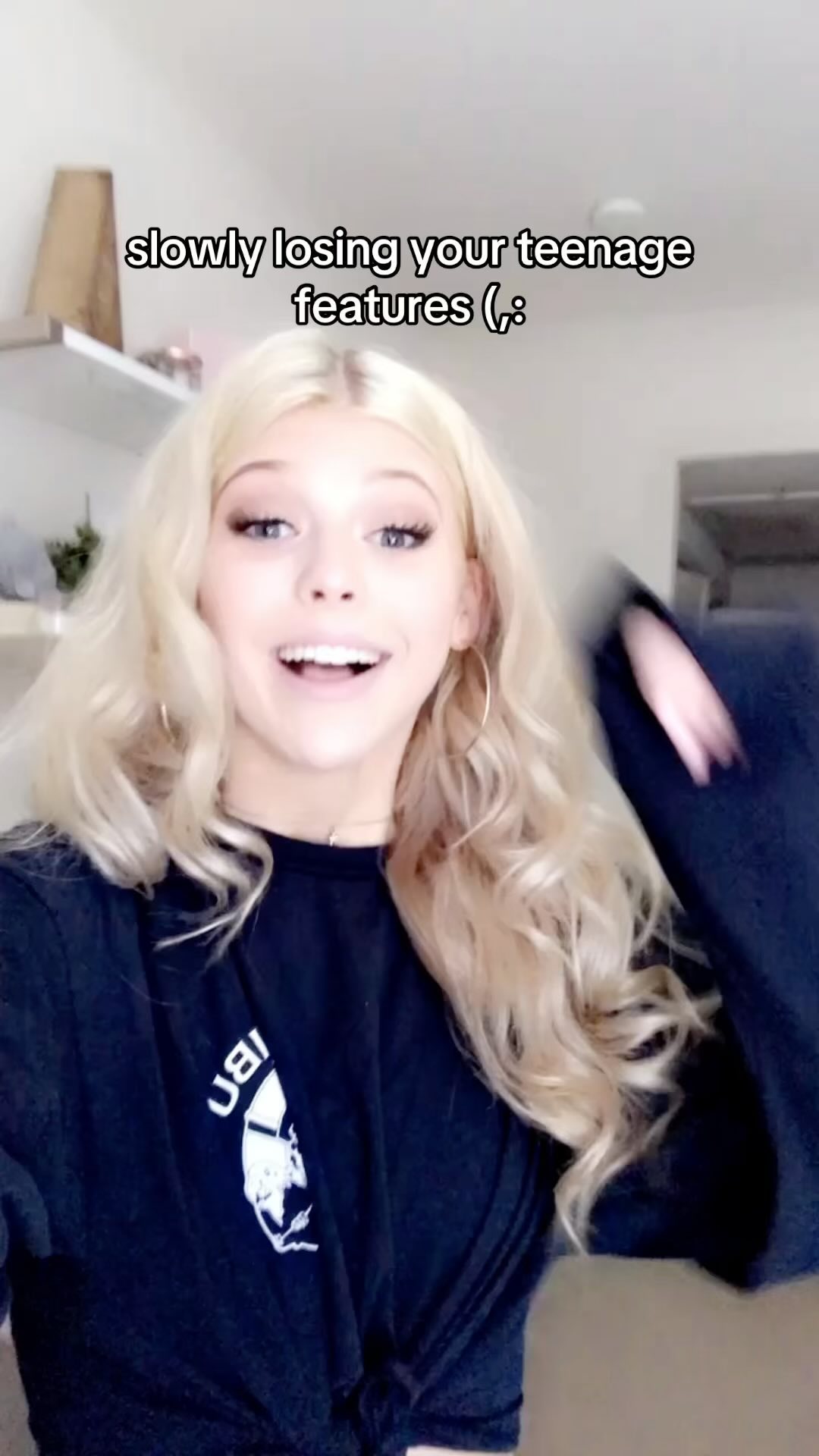 a young woman with blonde, wavy hair and hoop earrings stands indoors, wearing a black graphic tshirt. the text "slowly losing your teenage features ;_;" is superimposed on the image. a shelf with items can be seen in the background.