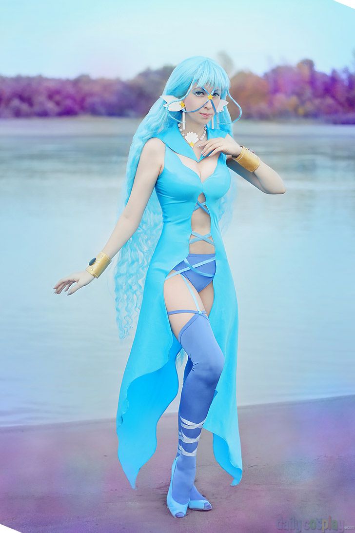 a woman in a blue anime cosplay costume stands on a beach near the water. she has long blue hair and is wearing a light blue dress with cutouts, matching thighhigh boots, and gold arm cuffs. she also has a necklace and a headpiece with flowerlike details. the background shows a blurred landscape with trees and water.