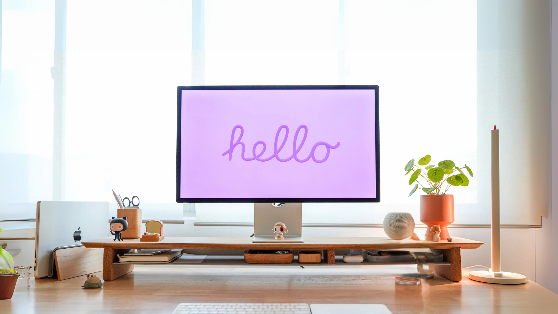 A desktop workspace with monitor displaying 'hello' in purple, wooden monitor stand, small potted plants, and minimalist office accessories