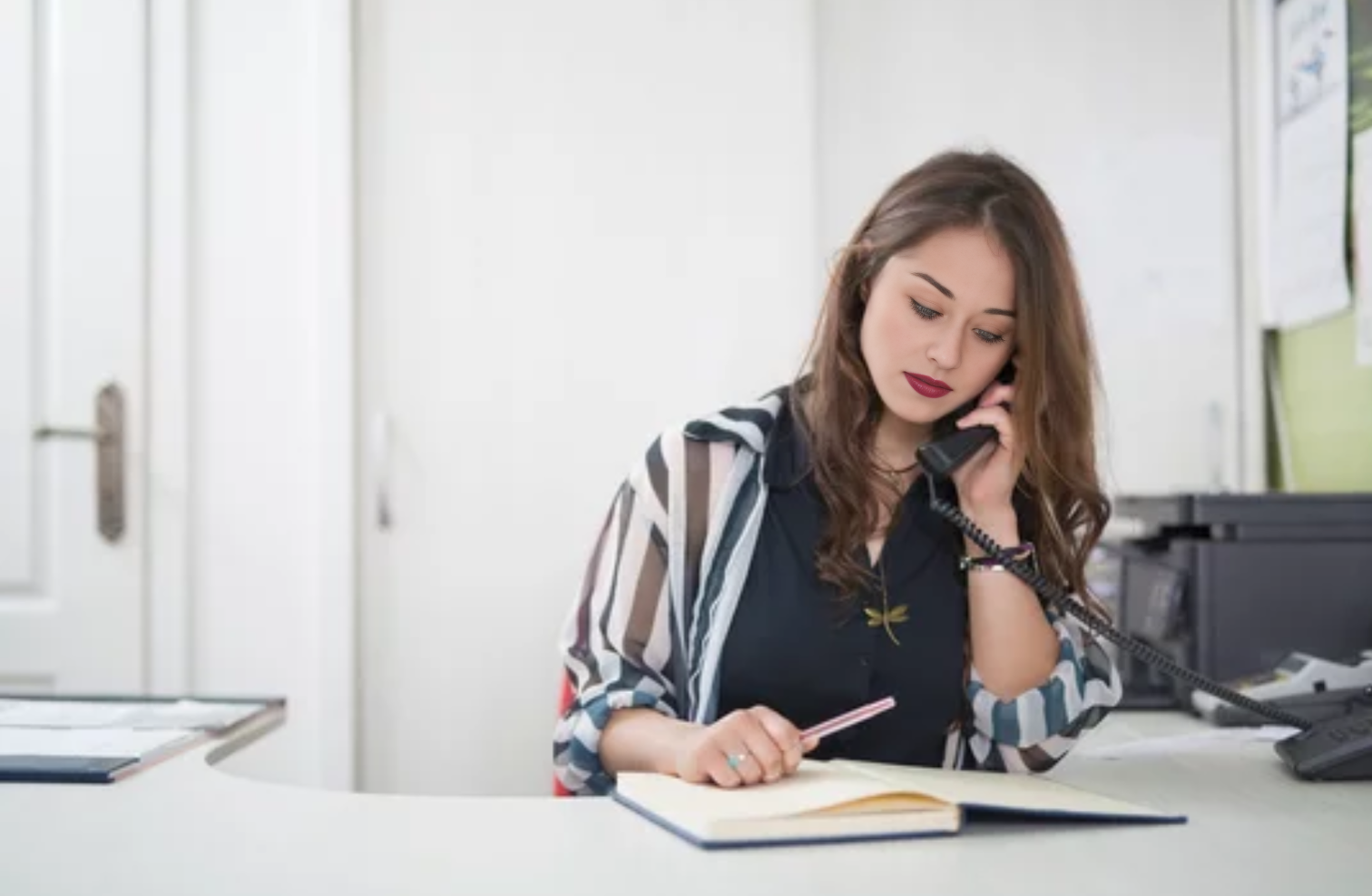 Professional taking notes while speaking on phone in bright office setting, wearing business attire