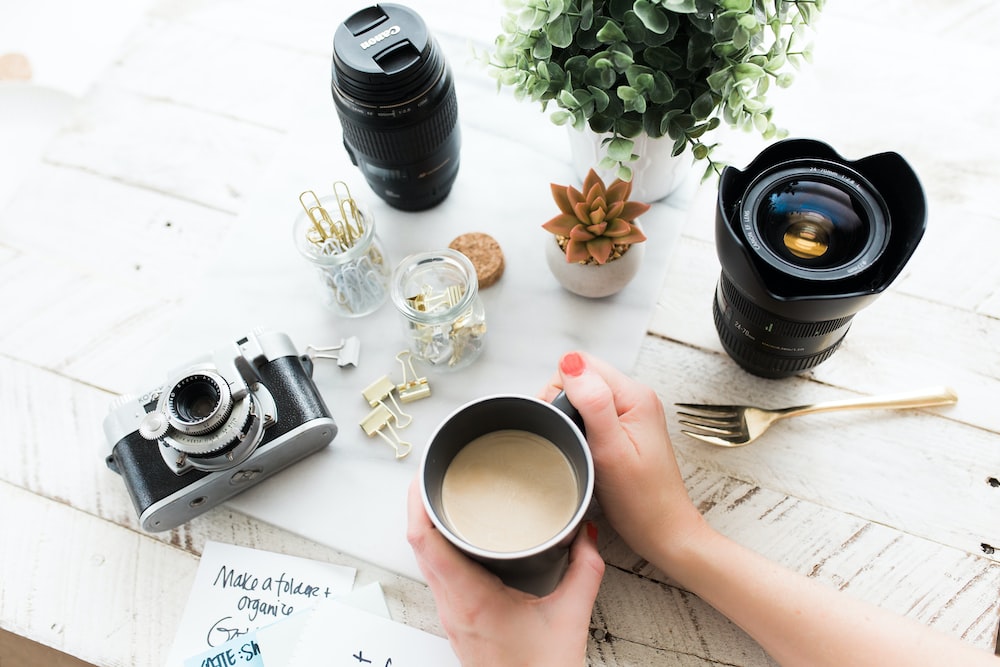 pessoa segurando uma caneca de café de cerâmica preta ao lado de uma câmera DSLR preta