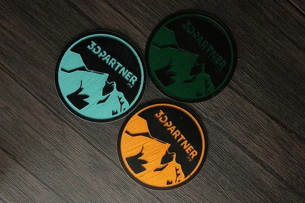 Tres posavasos impresos en 3D con logo de 3DPartner en turquesa, verde y naranja, con perímetros visibles