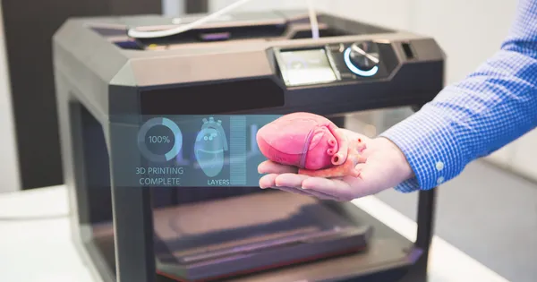 Mano sosteniendo un modelo de corazón impreso en 3D frente a una impresora, aplicación médica