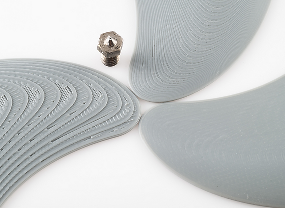 Piezas grises impresas en 3D junto a una boquilla, mostrando el material depositado capa por capa