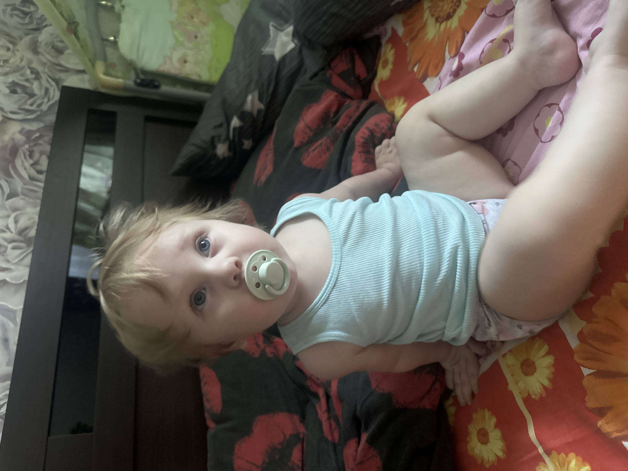 Infant resting on a bed in Sayanogorsk, bottle and pacifier nearby.