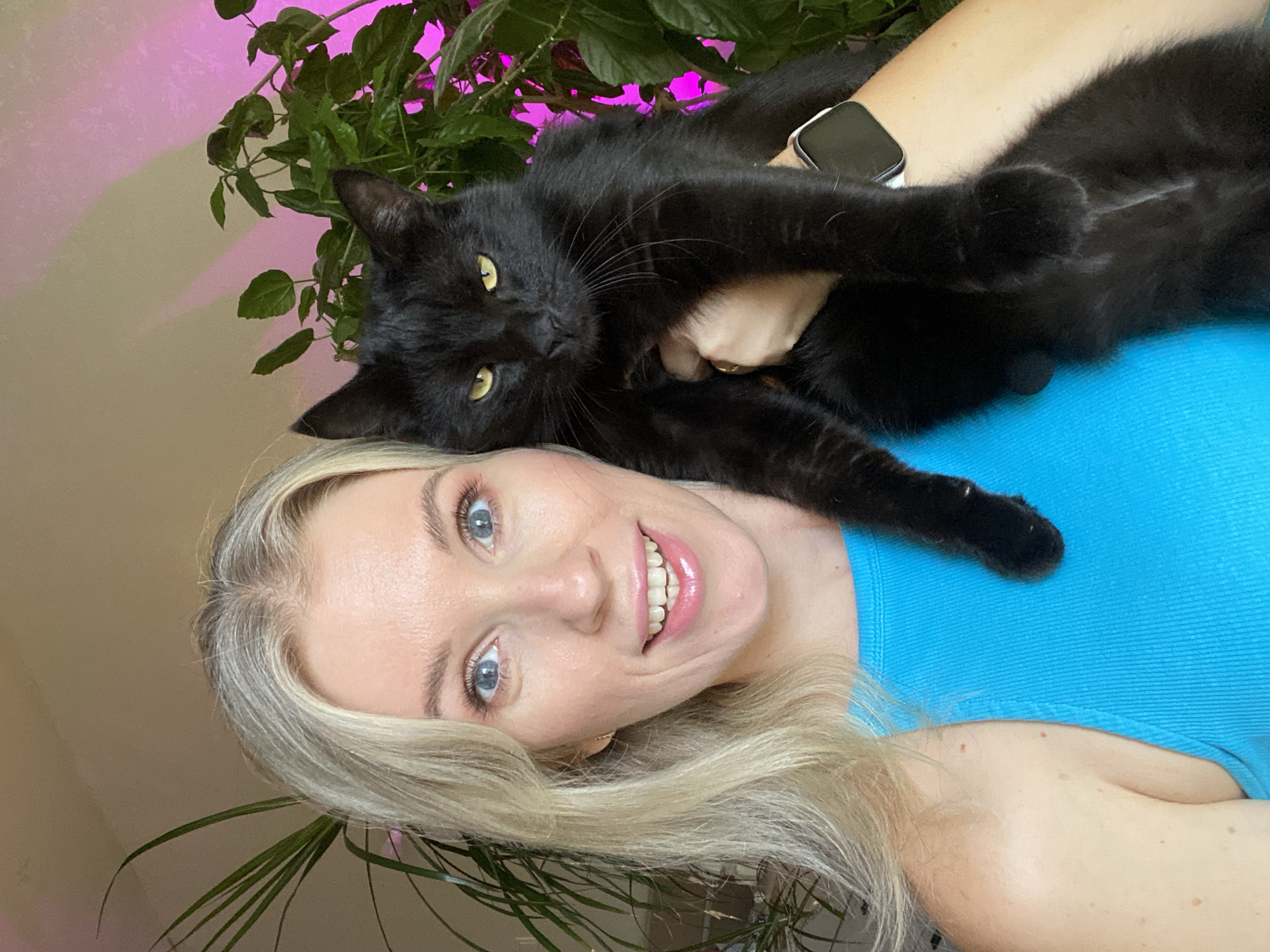 Holding a black cat on camera, mic clipped, blue tank top.