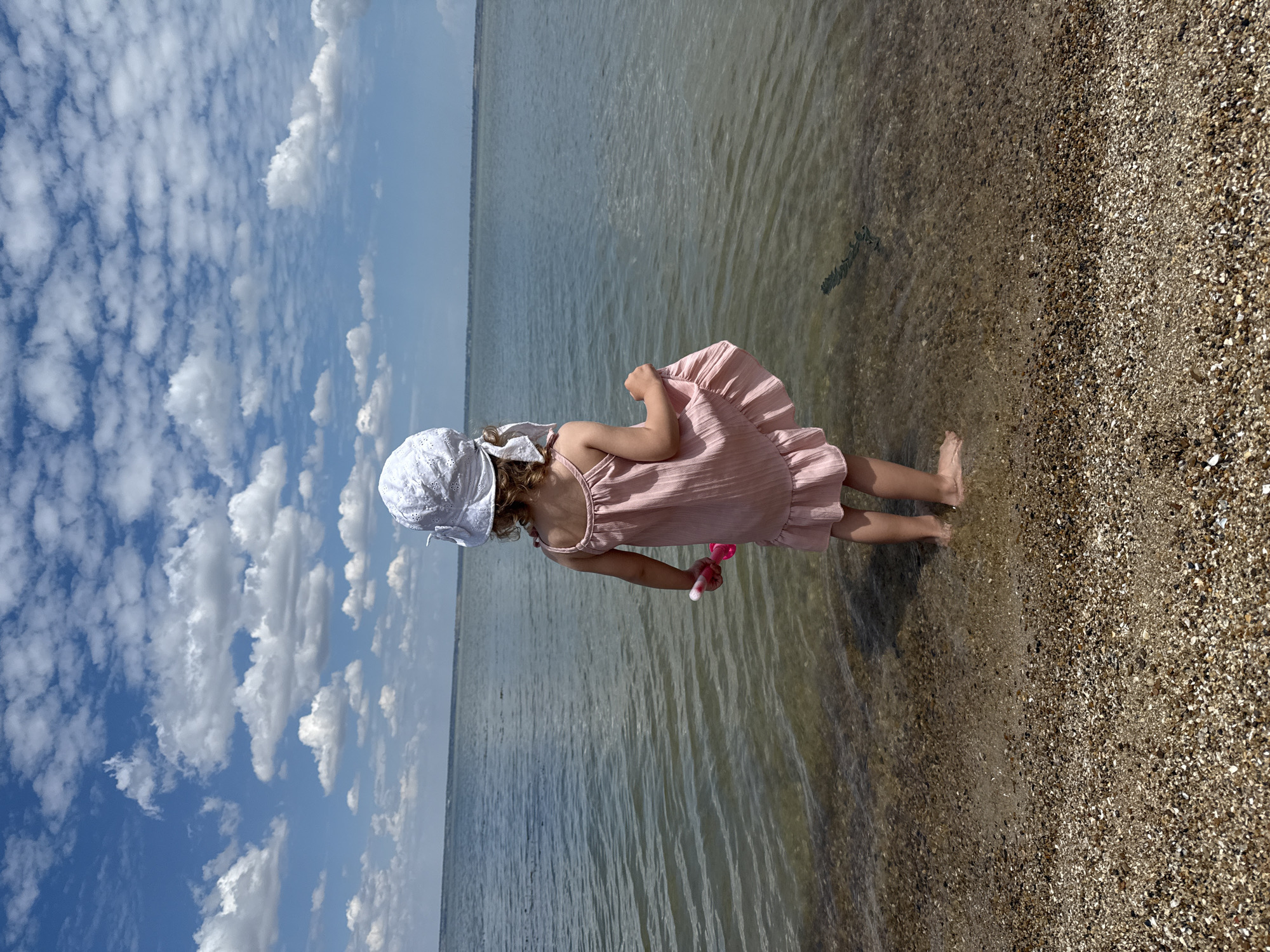 Small child at water's edge, pink dress and toy in hand
