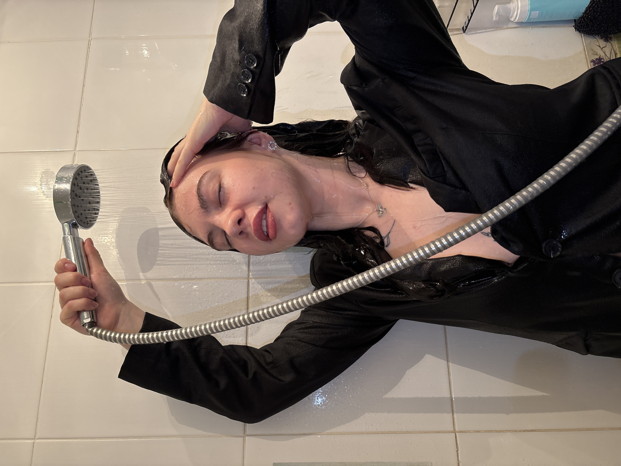 Woman in blazer lies on bathroom floor under shower