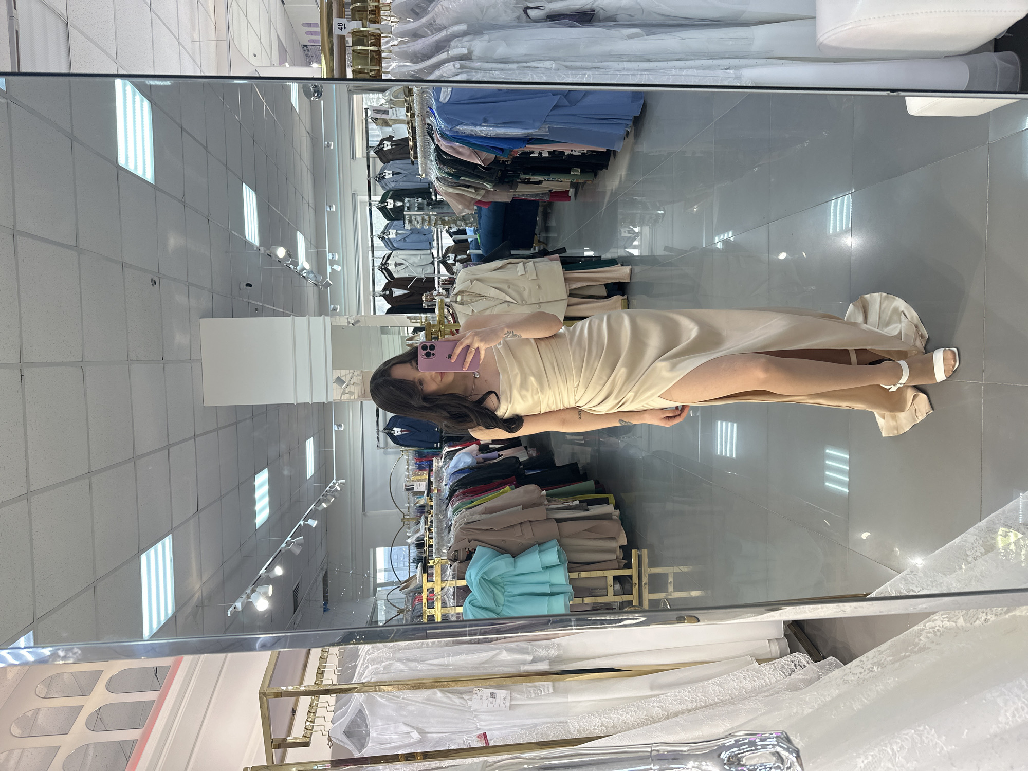 Woman takes mirror selfie in beige dress with pink phone