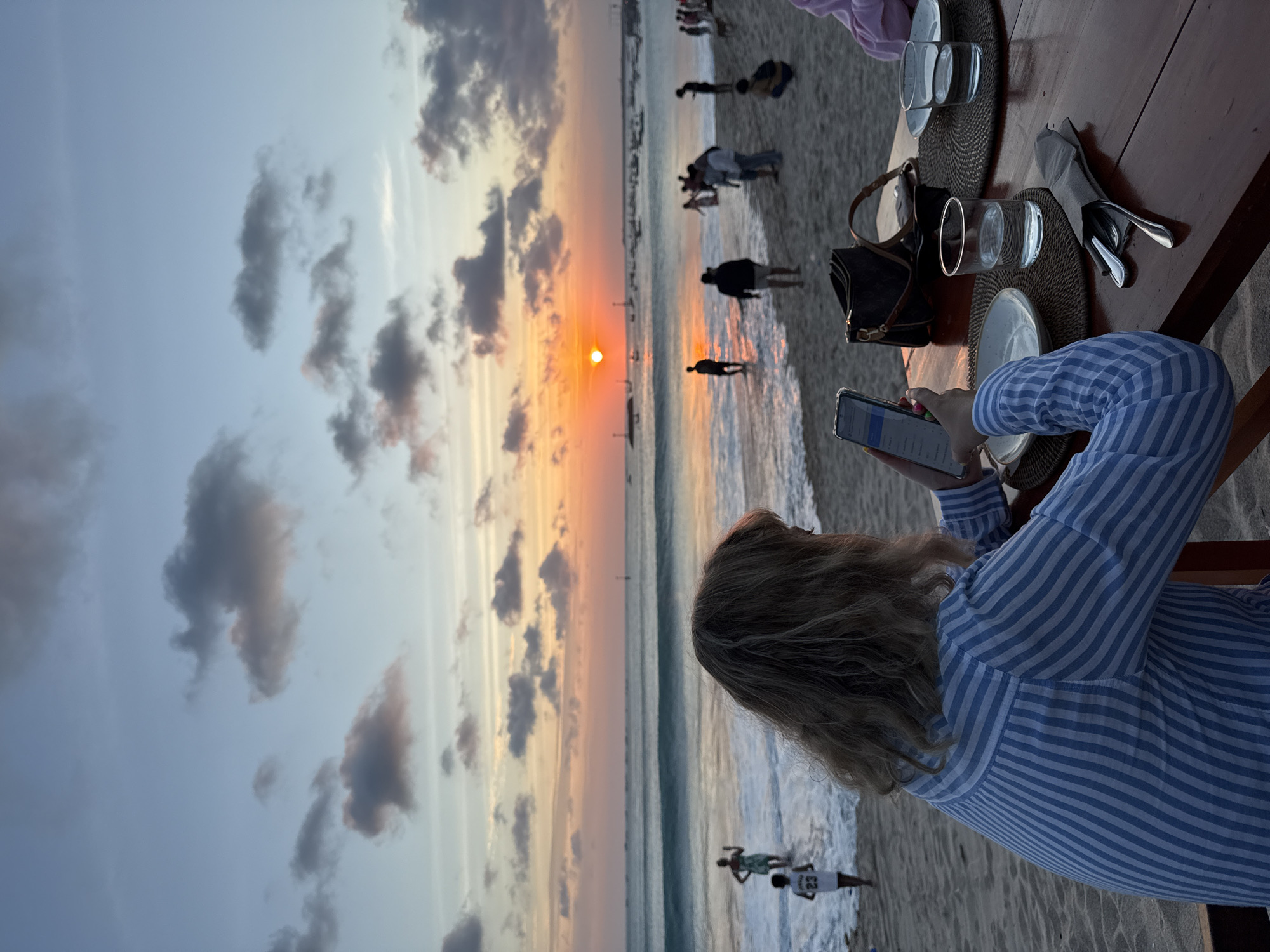 Woman uses phone at beach table during Kuta sunset