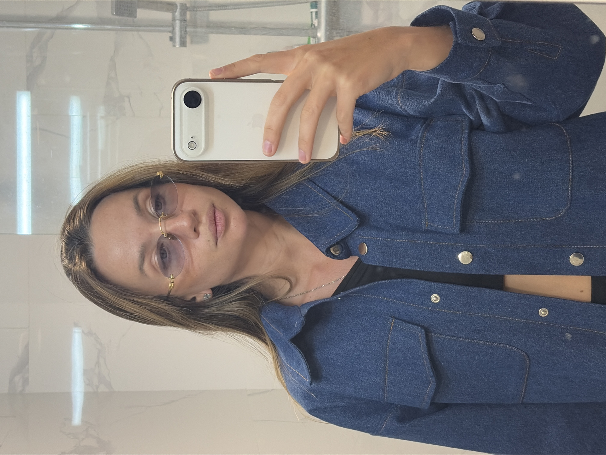 Woman takes bathroom selfie wearing denim jacket and glasses