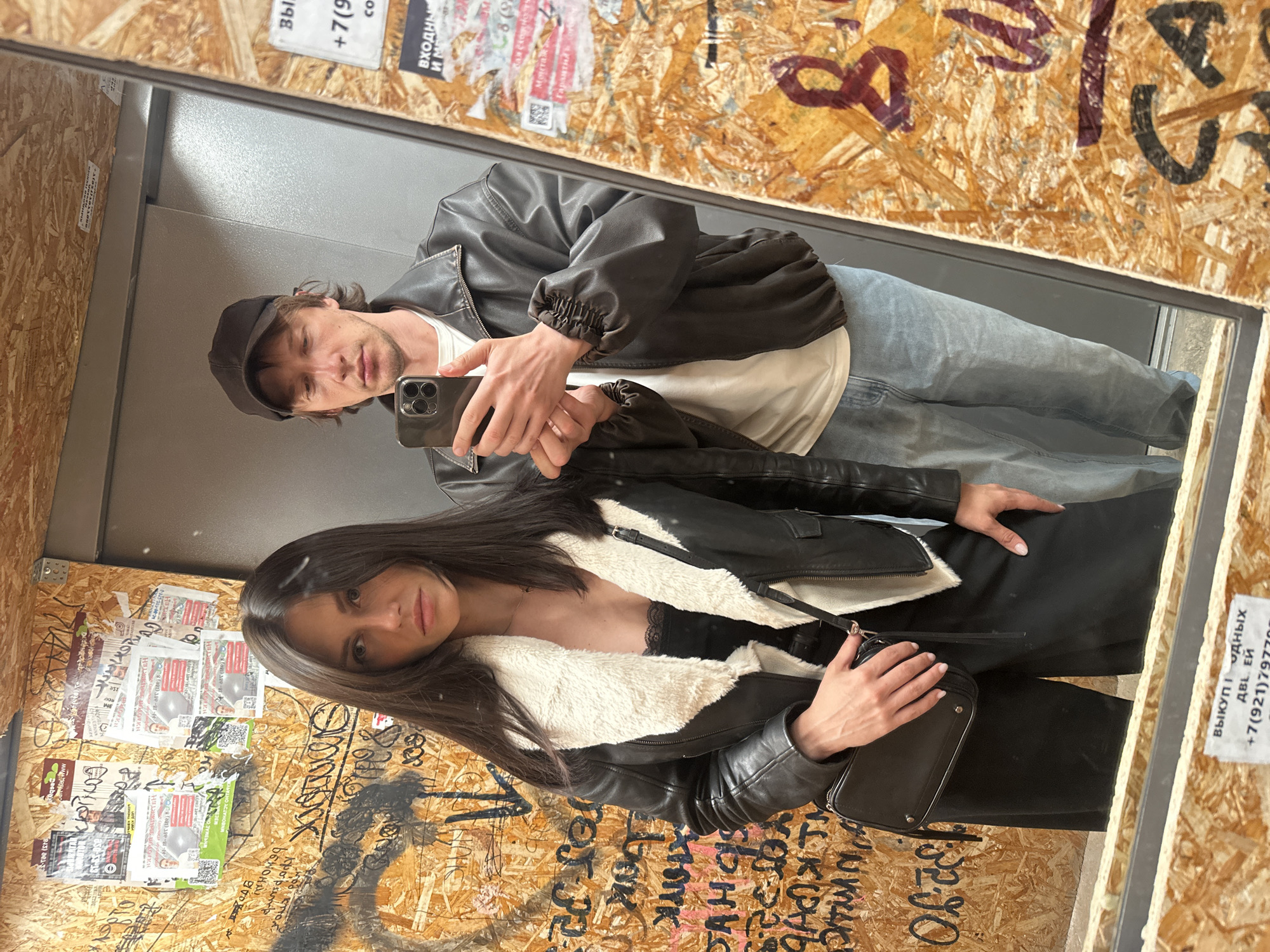 Couple takes elevator mirror selfie, woman in leather jacket