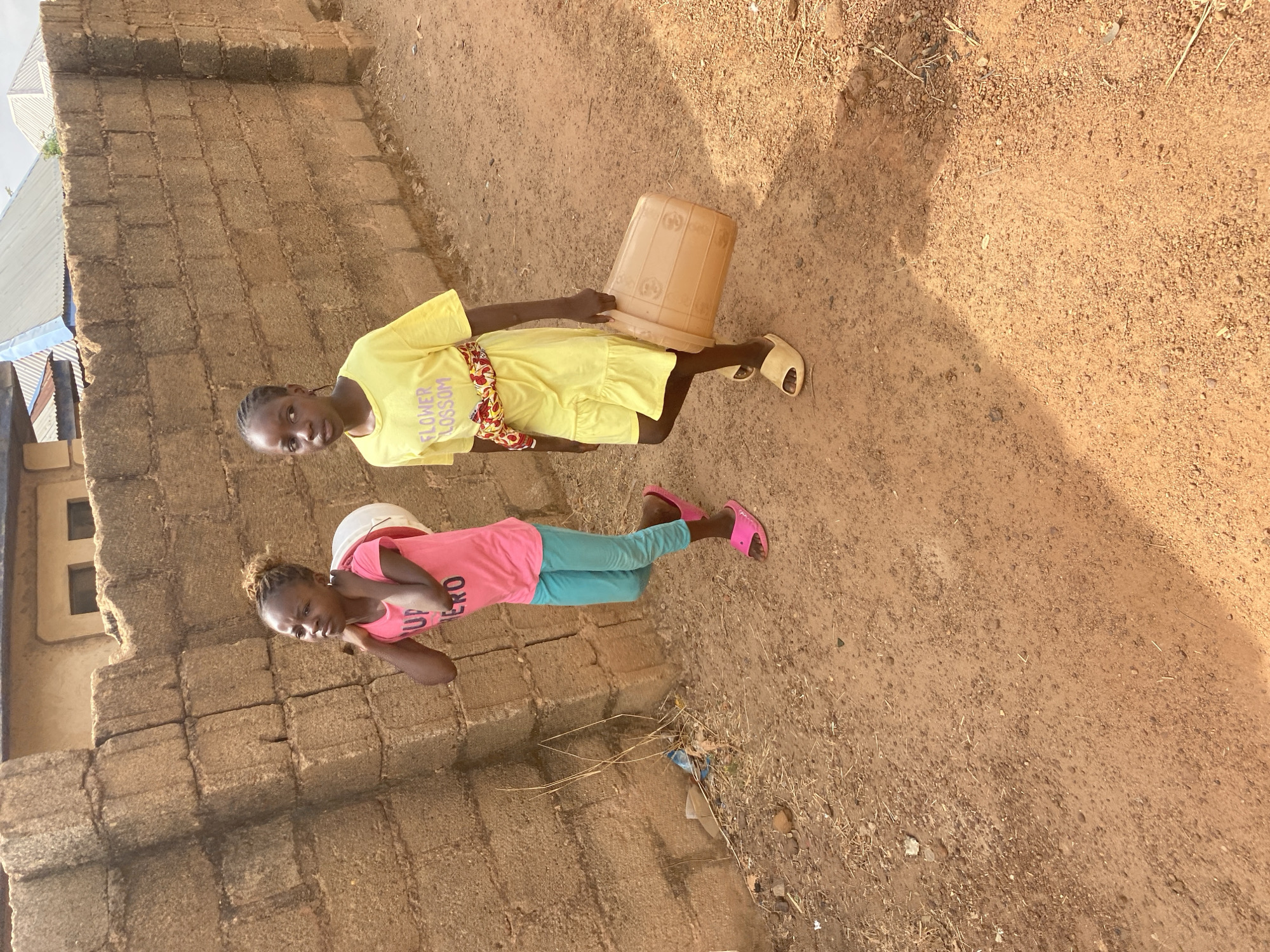 Two girls with bucket observed on dirt path in Bukuru