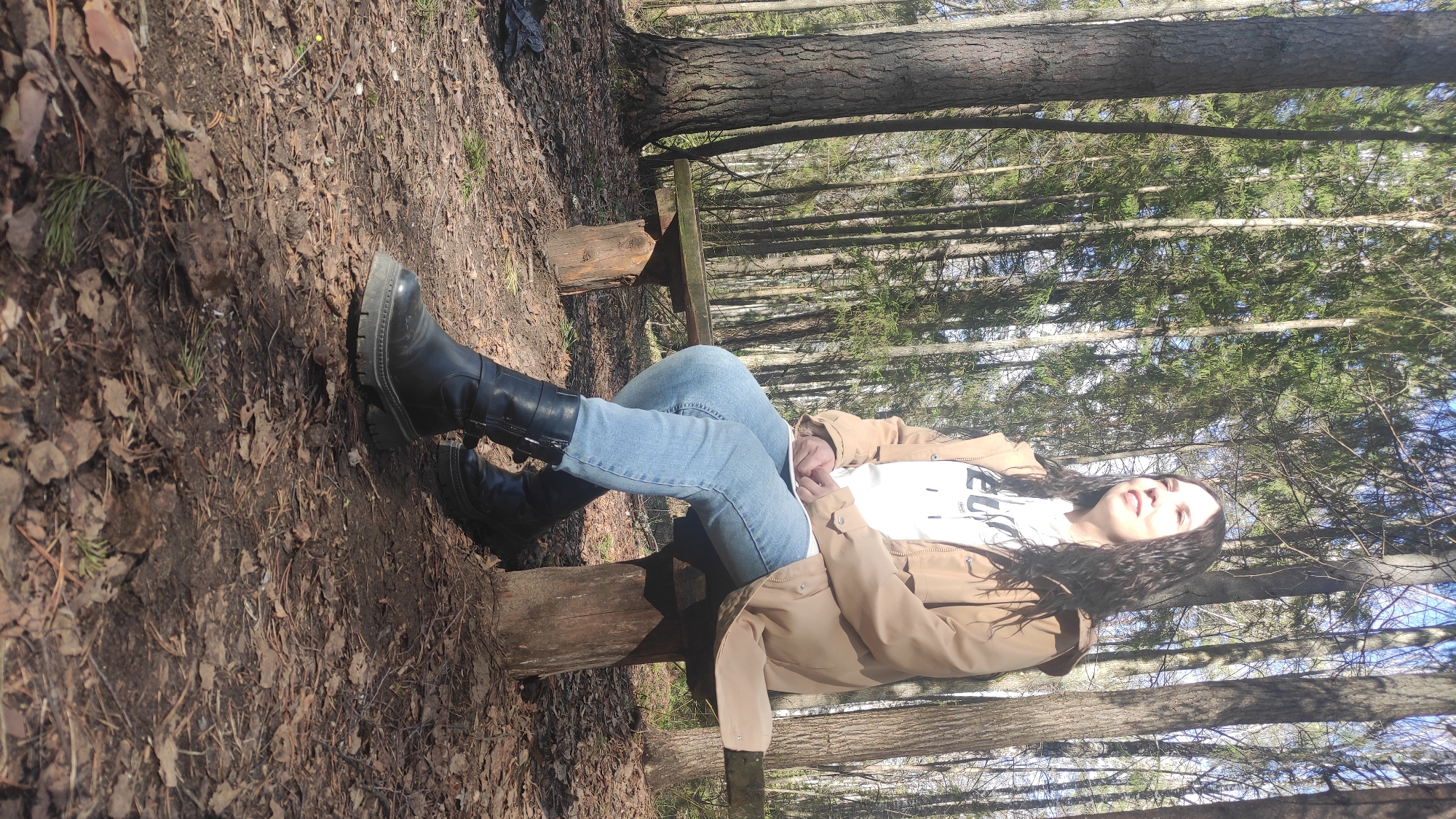 Woman in tan jacket sits on wooden bench in forest