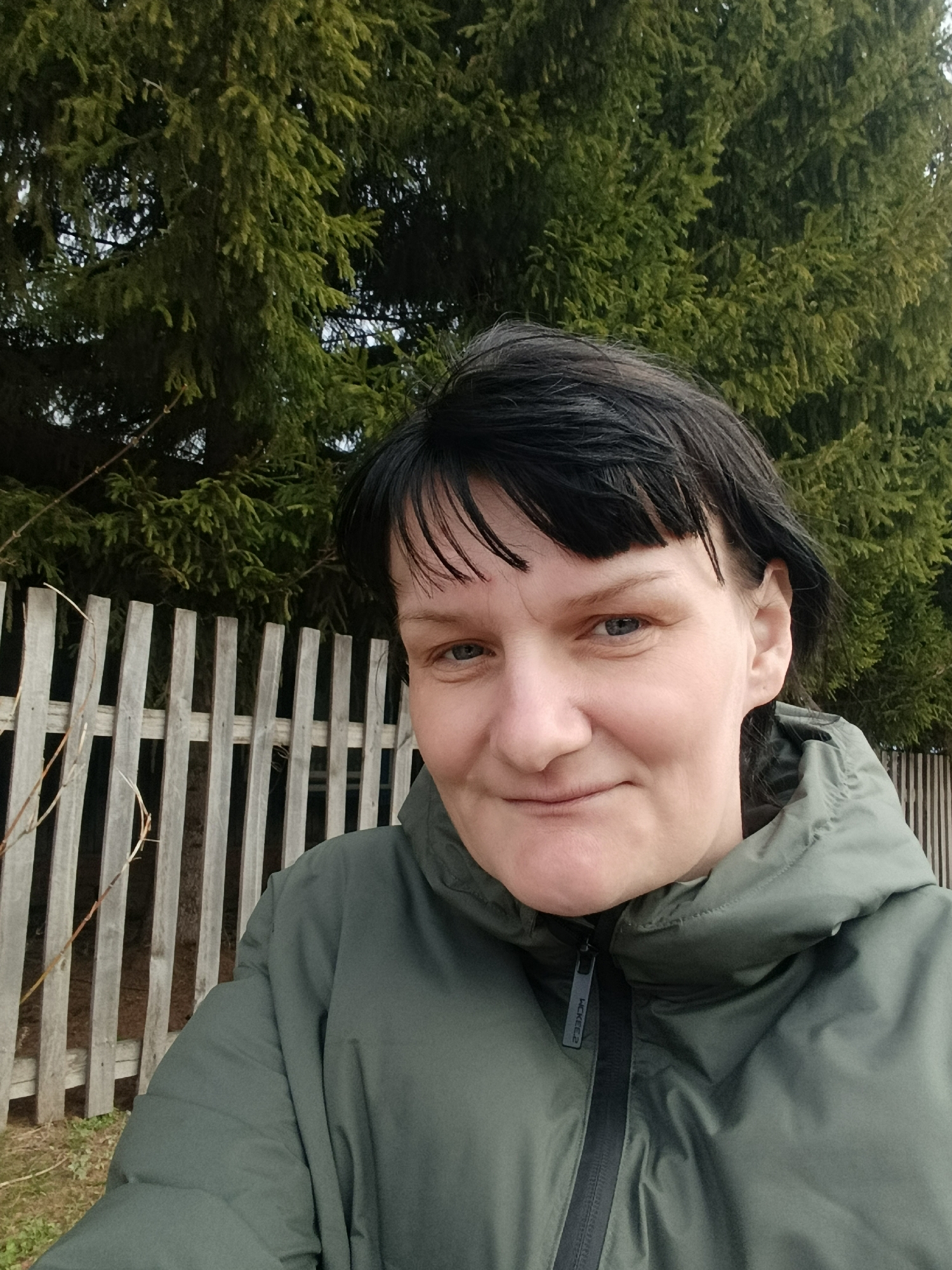 Woman in green jacket observed near wooden fence, trees