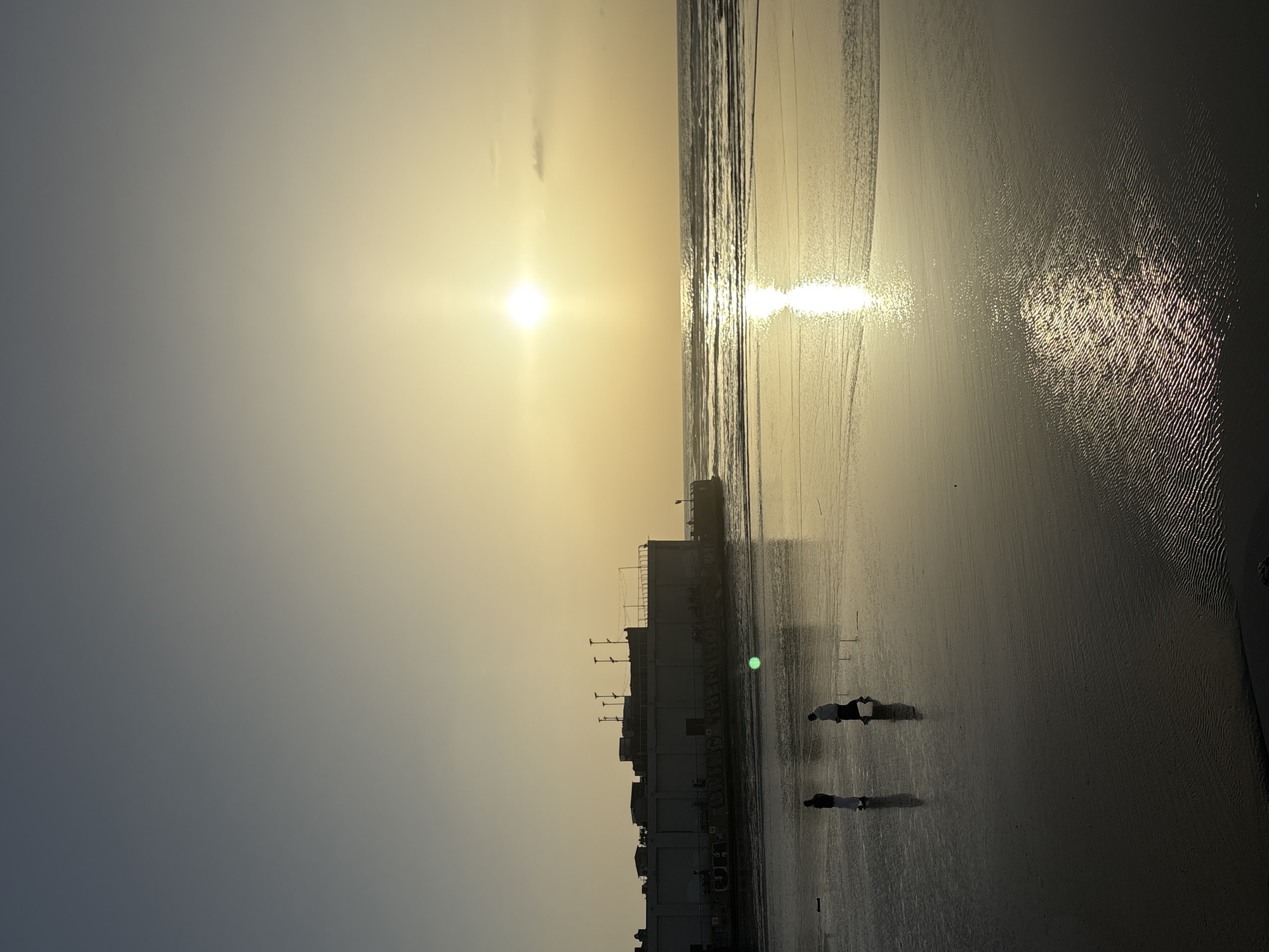 Two people walk on beach at sunset near shoreline building