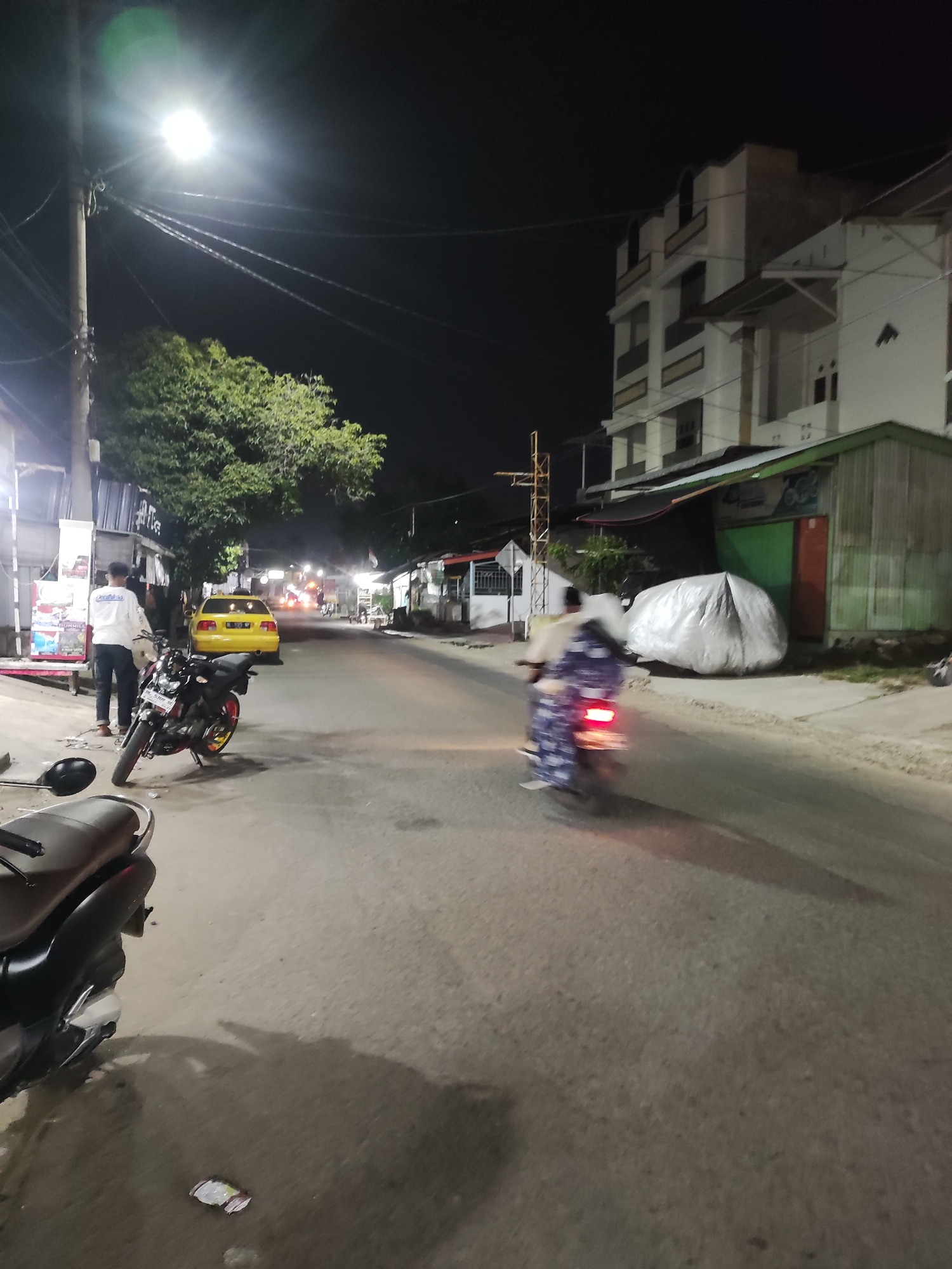 Man stands beside motorcycle on lit Lhokseumawe street
