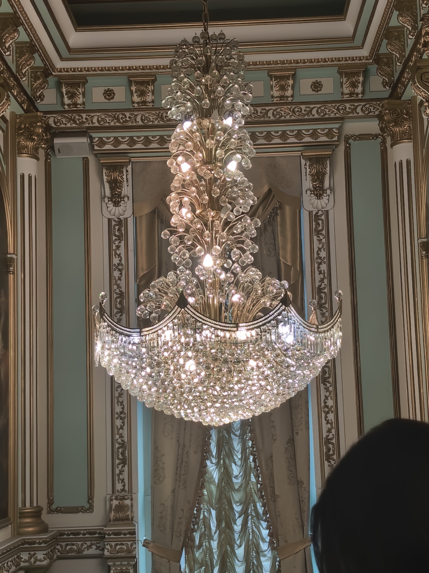 Inside a gilded ballroom or palace, chandeliers and classical murals throughout