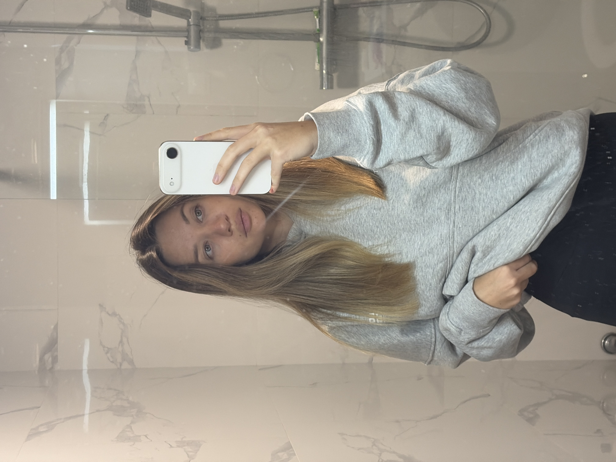 Woman takes bathroom mirror selfie in grey sweatshirt