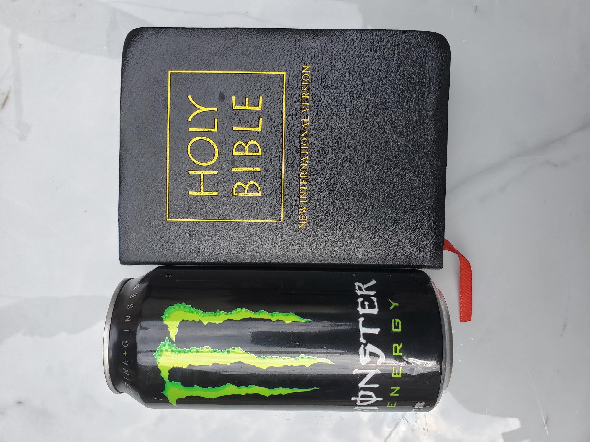 Monster Energy can photographed beside Bible on marble surface