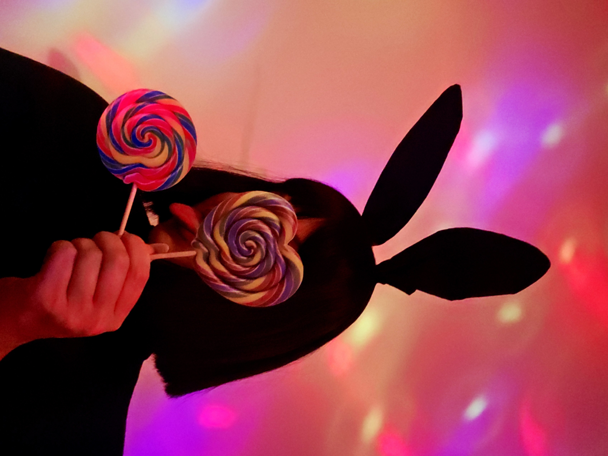 Person in bunny ears holds lollipops under colorful lights