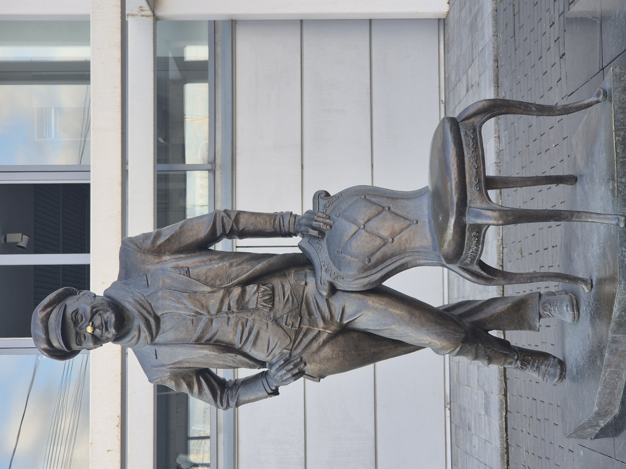 Bronze suited figure leans casually against an ornate outdoor chair.