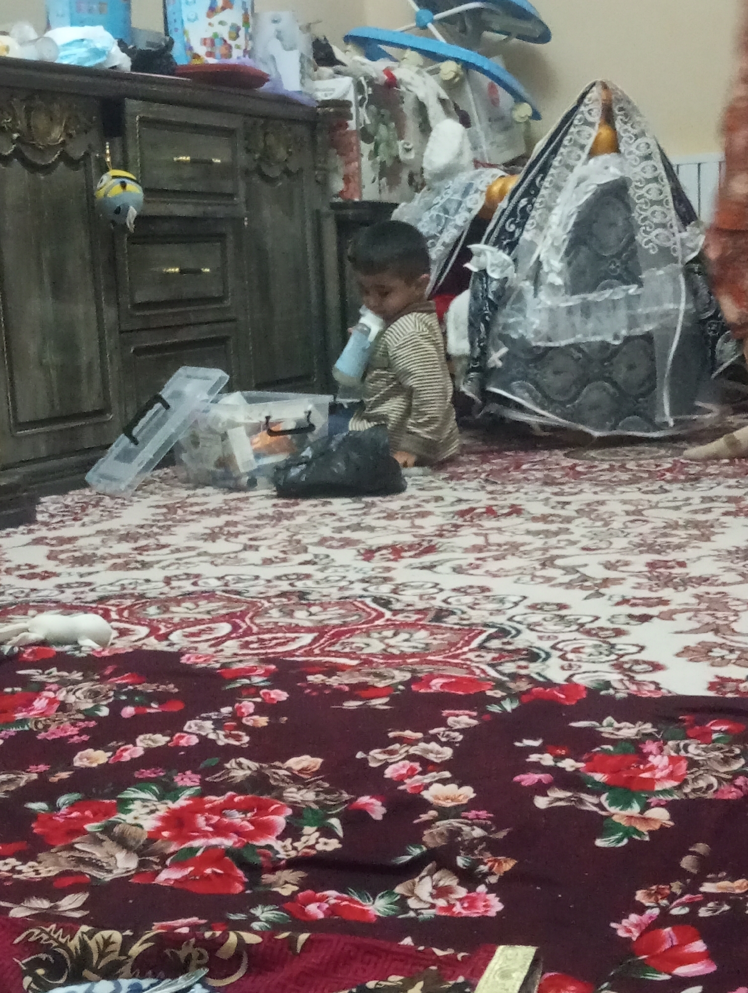 Child drinks from bottle on rug near household items
